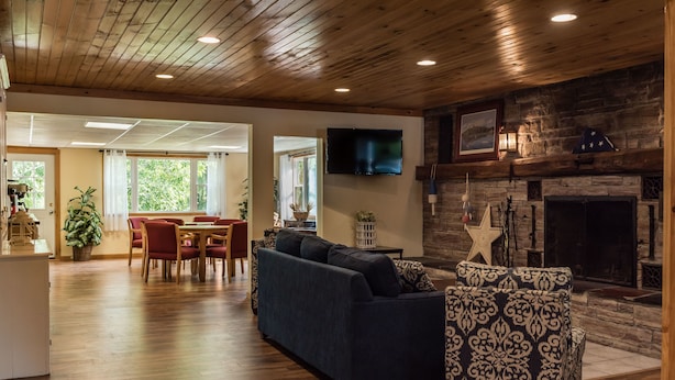 A spacious living room with a wooden ceiling, a fireplace, a television, and a dining area with red chairs and a table. - Capt.'s Inn & Suites (Alexandria Bay)