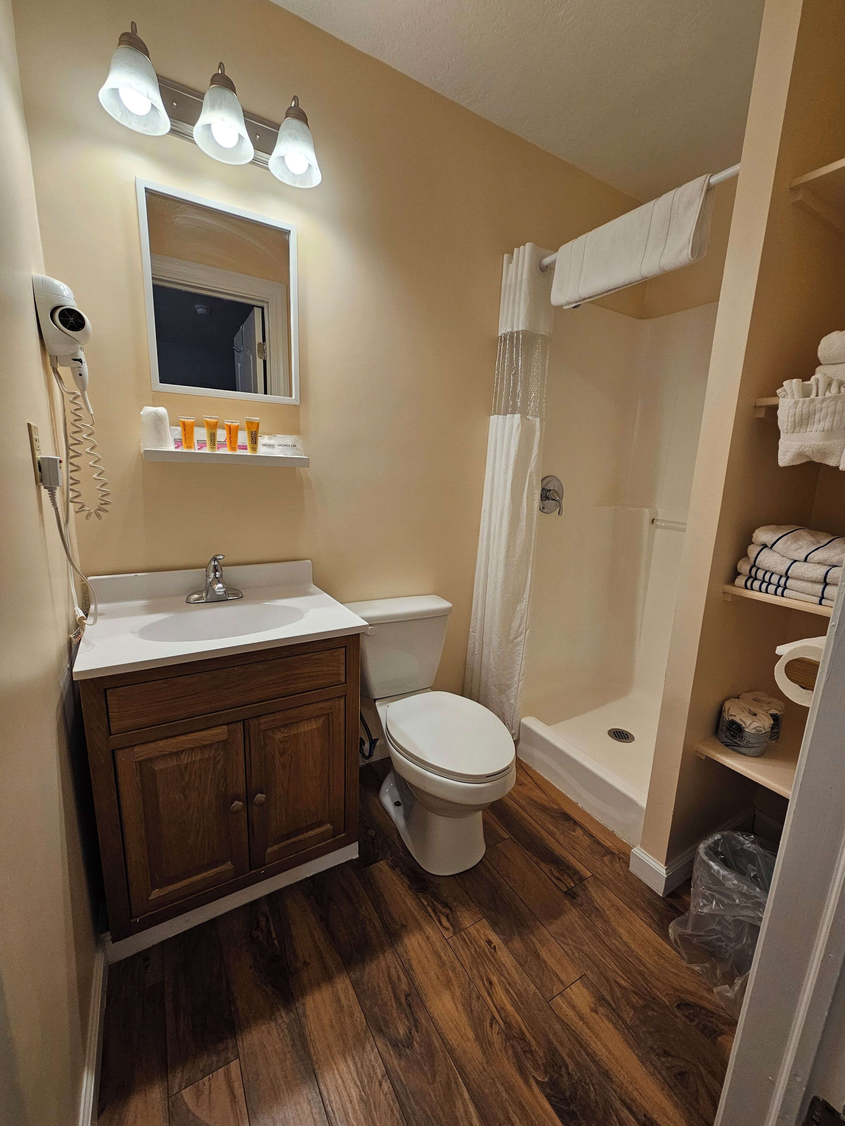 A bathroom with a wooden vanity, a toilet, a shower with a curtain, and a shelf with toiletries.