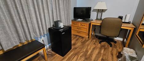 A small hotel room with a wooden floor, a desk with a computer, a chair, a TV, and a lamp.