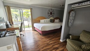A hotel room with a wooden floor, a bed with white bedding, a desk with a computer, a chair, and a view of the outdoors through a sliding door. - Capt.'s Inn & Suites (Alexandria Bay)
