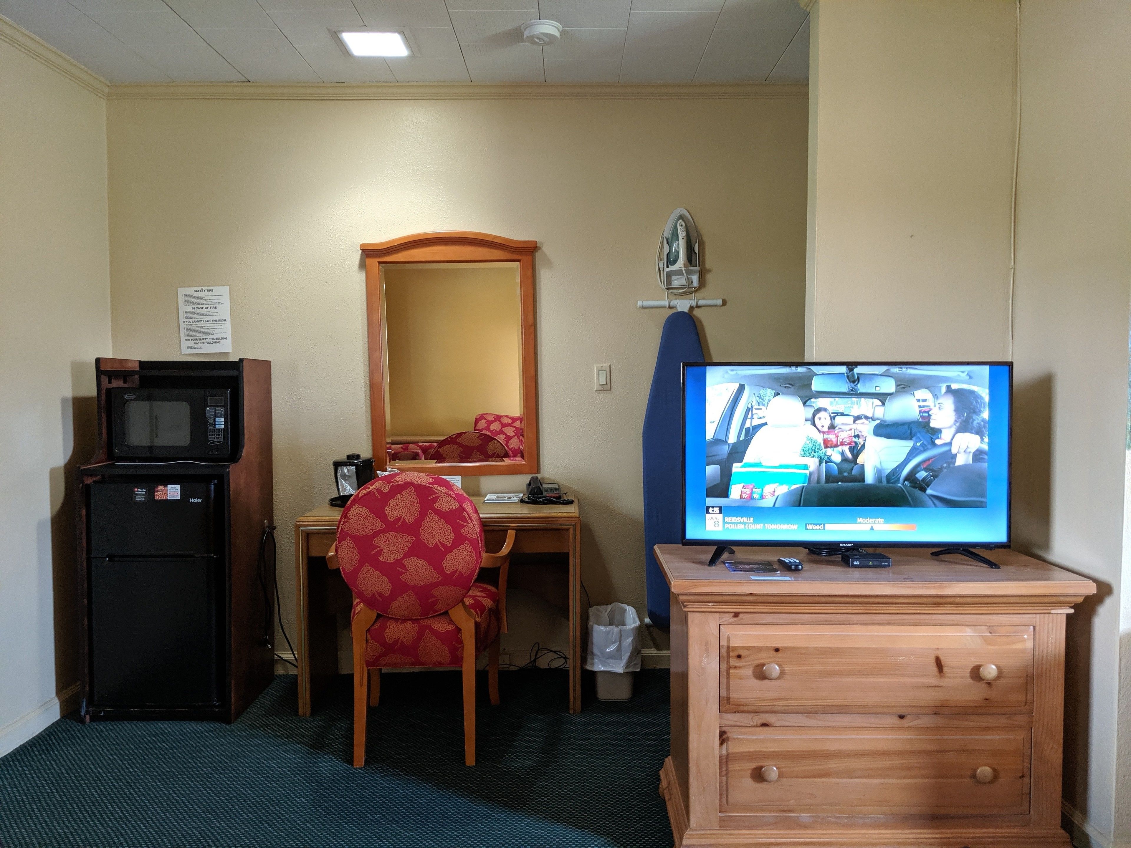 A hotel room with a wooden desk, a television, a microwave, a refrigerator, a chair, and a dresser.