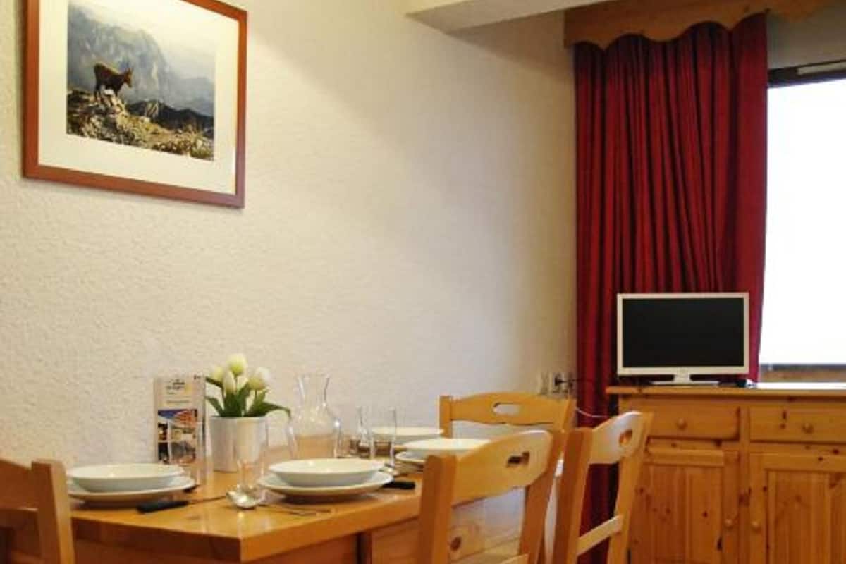 A wooden dining table with chairs, a framed picture on the wall, a television on a stand, and a window with red curtains.