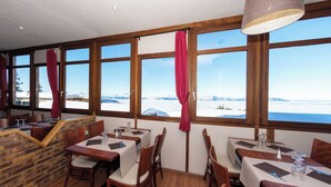 A restaurant with wooden tables and chairs, large windows with curtains, and a view of a snowy landscape. - La Bérangère (Chamrousse)