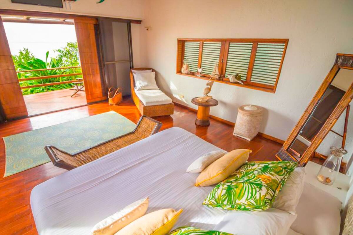 Une chambre à coucher avec un lit, des oreillers, une petite table, une chaise, une fenêtre avec des stores et un balcon.