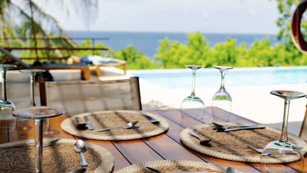 Mesa de comedor con cubiertos y cristalería, con vista a una piscina y follaje.