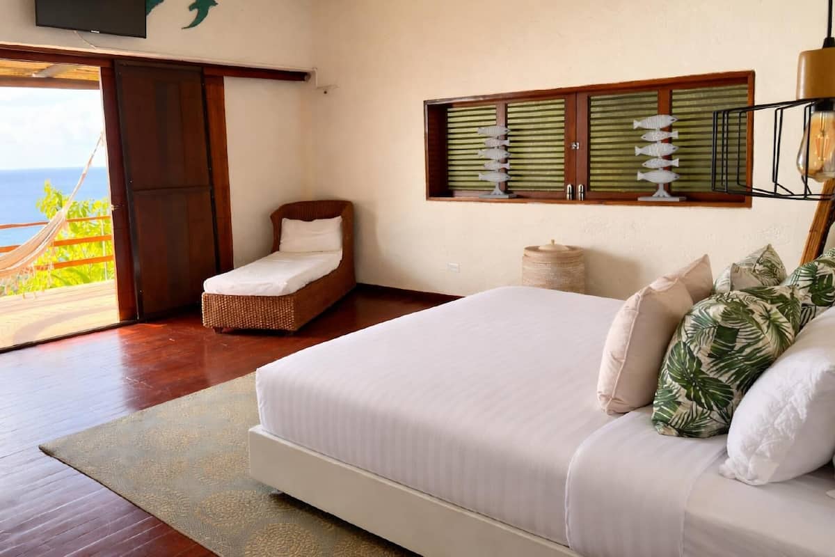 A bedroom with a bed, a small sitting area, a view of the ocean, and a hammock outside.
