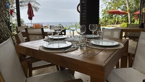 Mesa de madera para cuatro personas, con platos, cubiertos y vasos, con vistas a una piscina y palmeras.