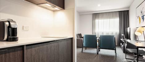 Habitación de hotel moderna con kitchenette, zona de estar con sillas, escritorio con lámpara y un ventanal con cortinas.