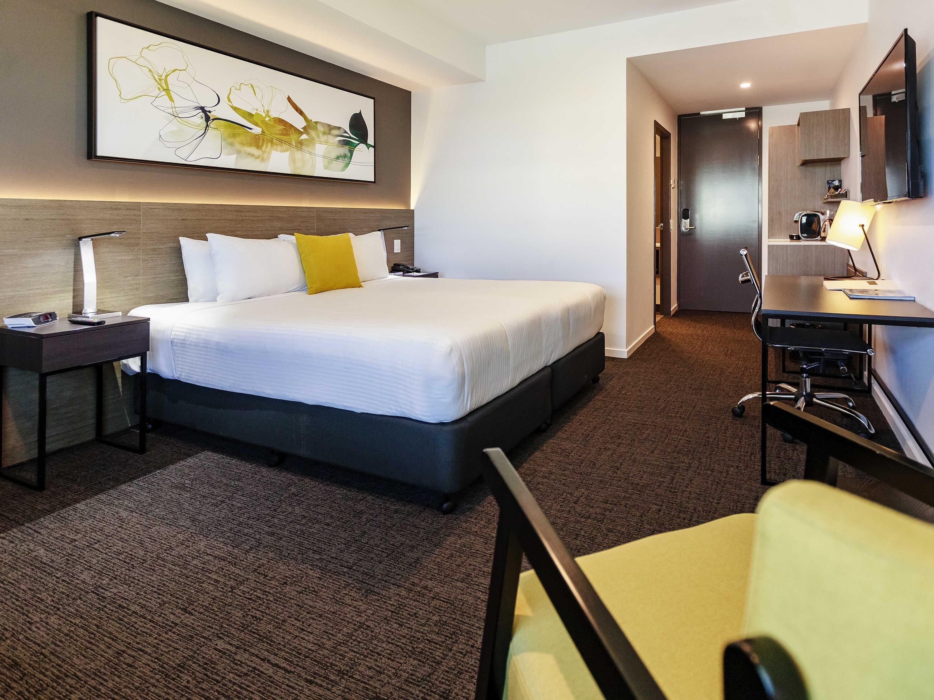 Ein Hotelzimmer mit Bett, Schreibtisch, Stuhl, Schreibtischlampe, Nachttisch, Wand-Fernseher und einem Gemälde.
