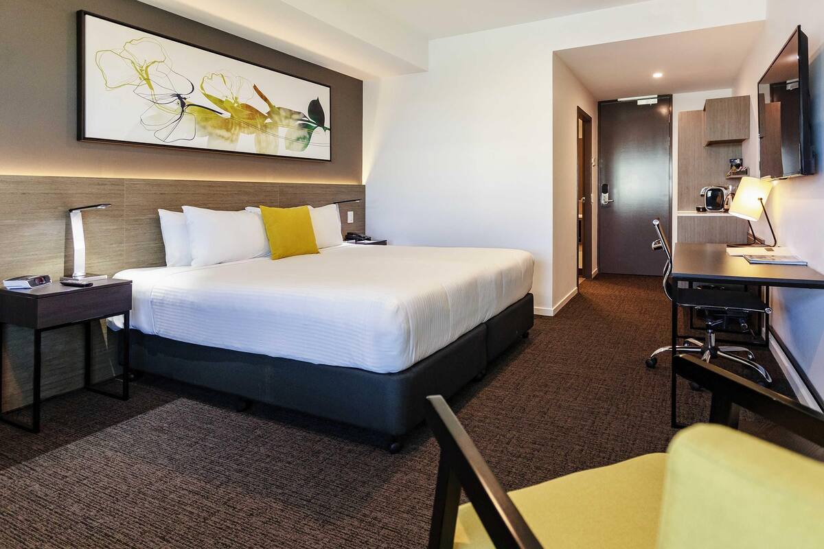A hotel room with a bed, a desk, a chair, a desk lamp, a nightstand, a wall-mounted TV, and a painting.