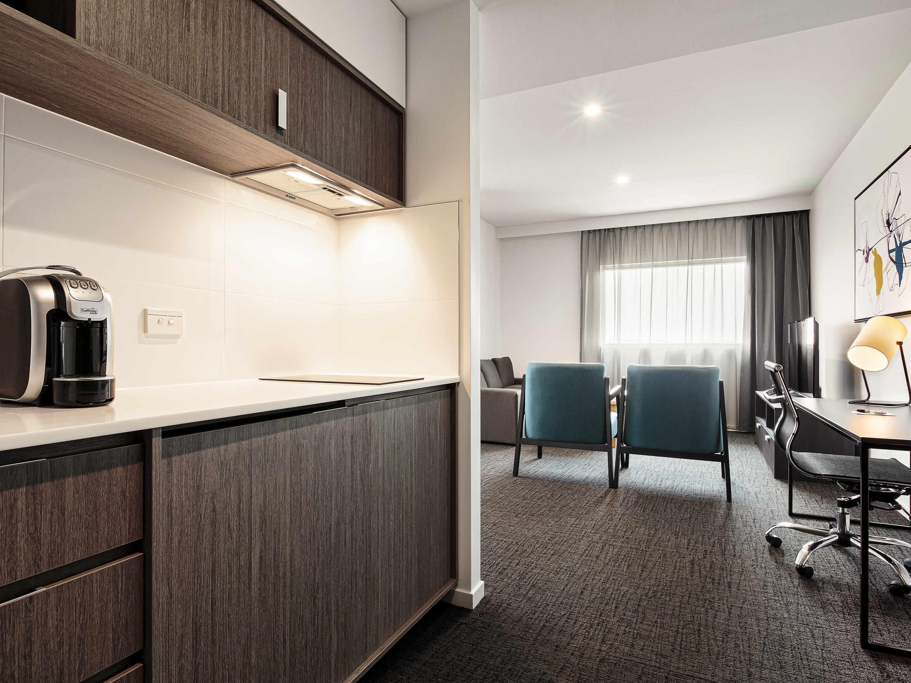 A modern hotel room with a kitchenette, a seating area with chairs, a desk with a lamp, and a large window with curtains.
