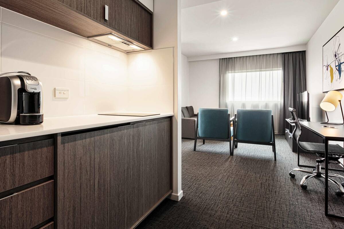 A modern hotel room with a kitchenette, a seating area with chairs, a desk with a lamp, and a large window with curtains.