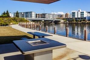 An outdoor patio with a grill and seating area overlooking a river, with buildings in the background. - Mercure Sunshine Coast Kawana Waters (Birtinya)