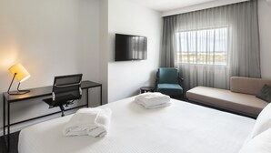 Una habitación de hotel con cama, escritorio con lámpara, silla, sofá, televisión y ventana con cortinas.