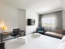 Une chambre d’hôtel avec un lit, un bureau avec une lampe, une chaise, un canapé, une télévision et une fenêtre avec des rideaux.