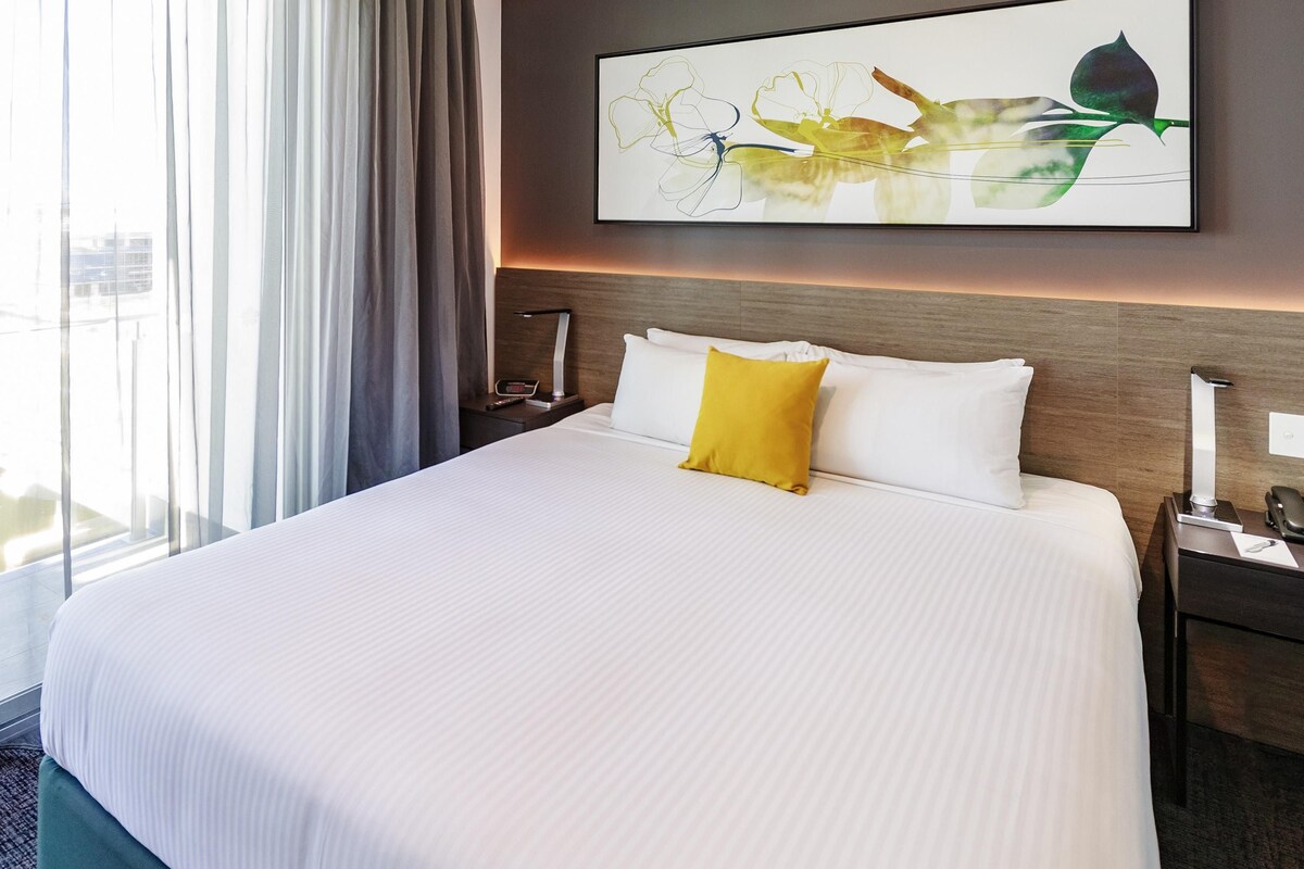 A hotel room with a large bed, a nightstand, a bedside table, a lamp, and a window with curtains.