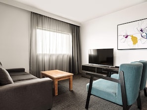 A modern hotel room with a flat-screen TV, a wooden coffee table, grey sofas, teal armchairs, and a large abstract painting on the wall. - Mercure Sunshine Coast Kawana Waters (Birtinya)