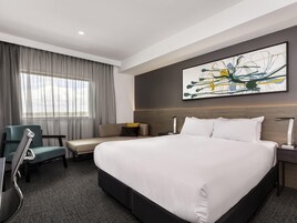 A hotel room with a large bed, a desk, a chair, a window with curtains, and a wall-mounted abstract painting.
