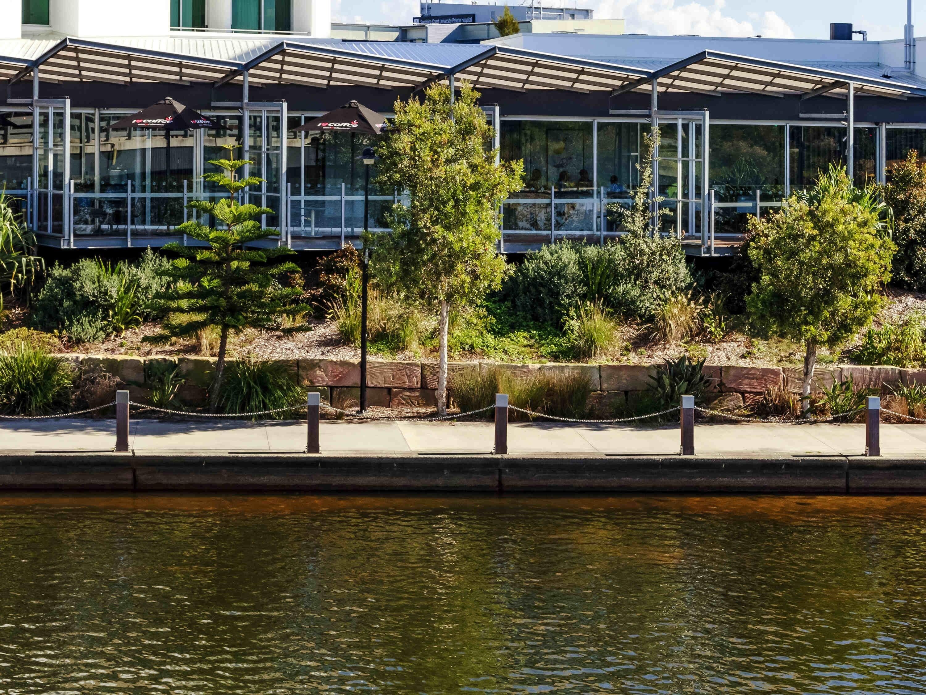 A modern building with a glass facade, a covered outdoor seating area, and a water feature in the foreground.