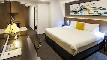 A hotel room with a large bed, a desk with a lamp, a nightstand, and a wall-mounted abstract painting.