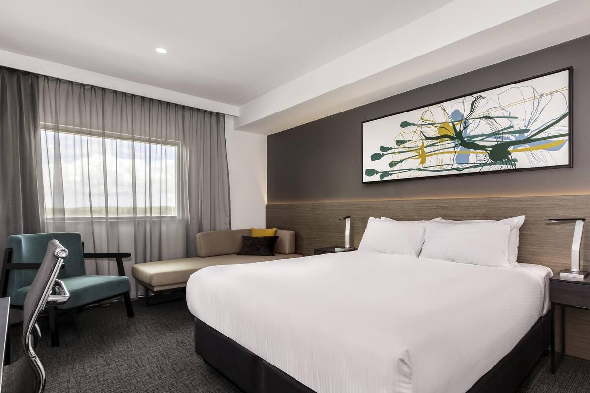 A hotel room with a large bed, a desk, a chair, a window with curtains, and a wall-mounted abstract painting.