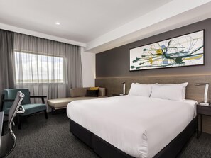 Une chambre d’hôtel avec un grand lit, un bureau, une chaise, une fenêtre avec des rideaux et un tableau abstrait accroché au mur.