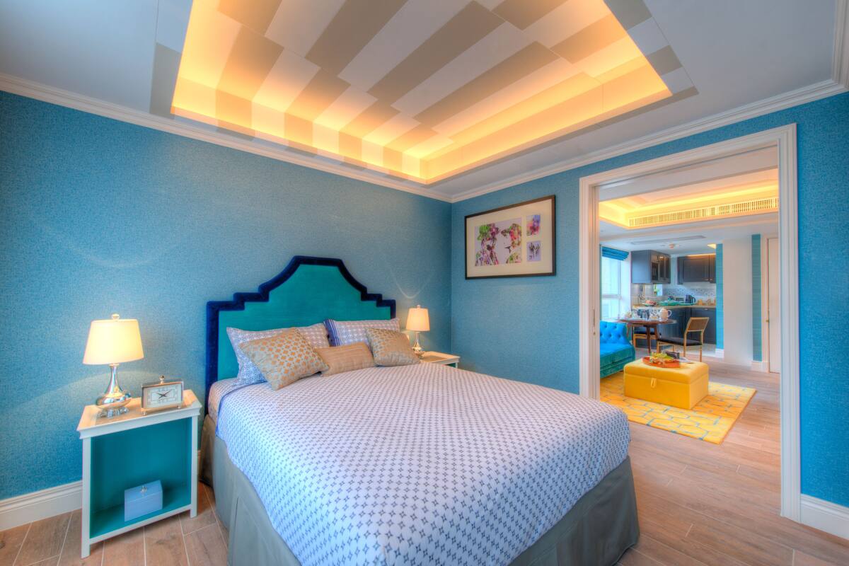 Un dormitorio con cama, mesitas de noche, pared azul, techo con iluminación empotrada y un acceso a una zona de cocina.