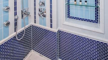 A bathroom with blue and white tiled walls, a shower with a handheld showerhead, and a window with shelves holding toiletries.