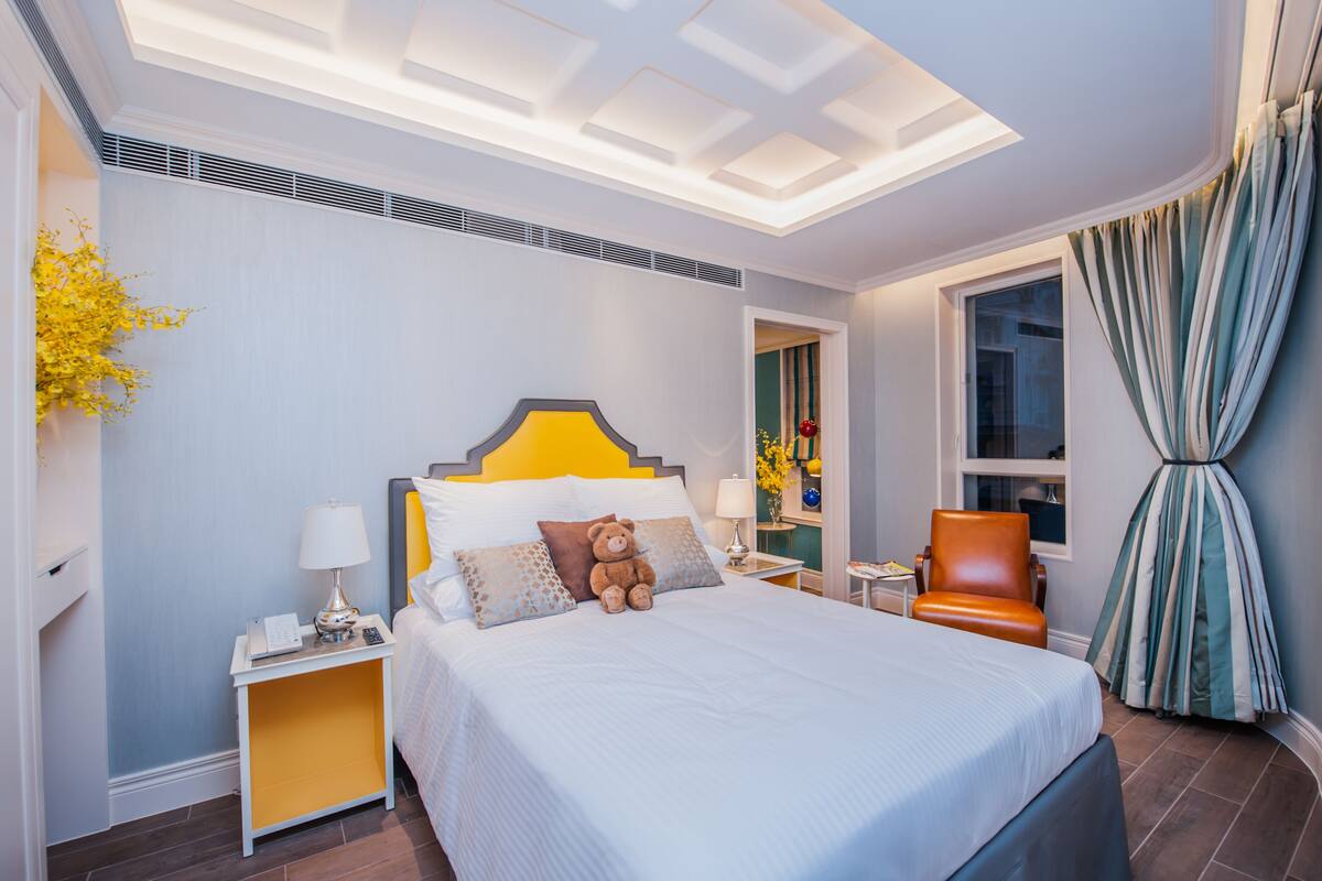 Una habitación de hotel con una cama grande, una mesita de noche con lámpara, una silla y una ventana con cortinas.