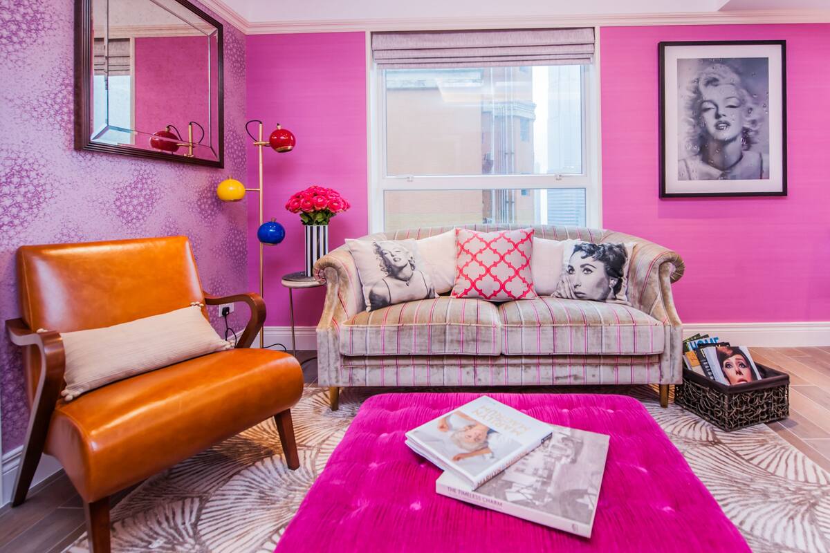Una sala de estar con pared rosa, un sofá con cojines estampados, un sillón de cuero, un espejo y una ventana con vistas.