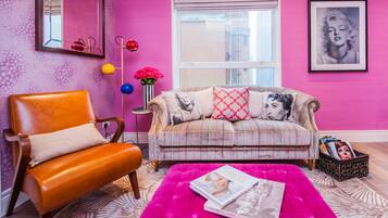 Ein Wohnzimmer mit einer rosa Wand, einem Sofa mit gemusterten Kissen, einem Ledersessel, einem Spiegel und einem Fenster mit Blick.