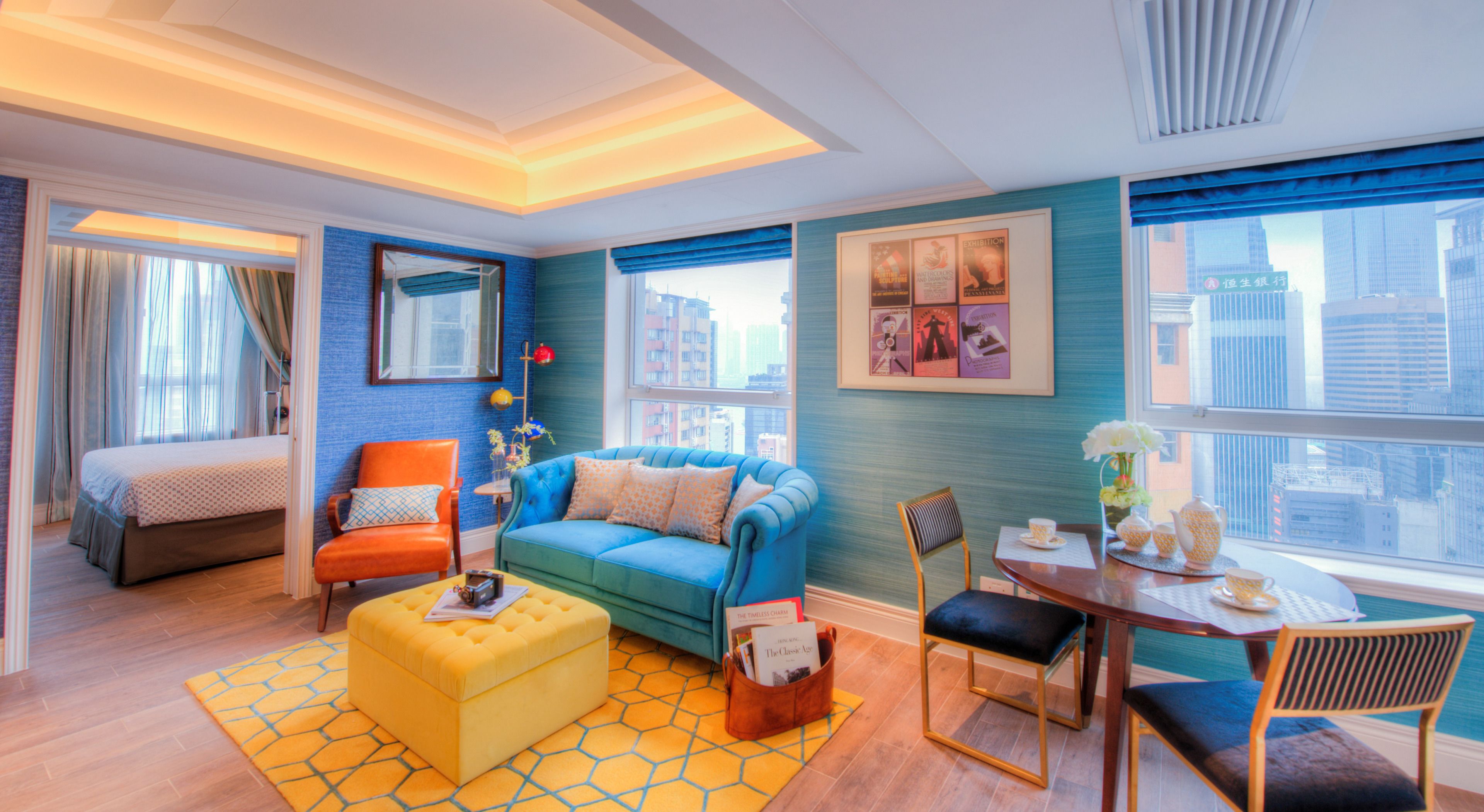 A modern hotel room with a large bed, a blue sofa, a yellow ottoman, a small table with a teapot, and a view of the city.