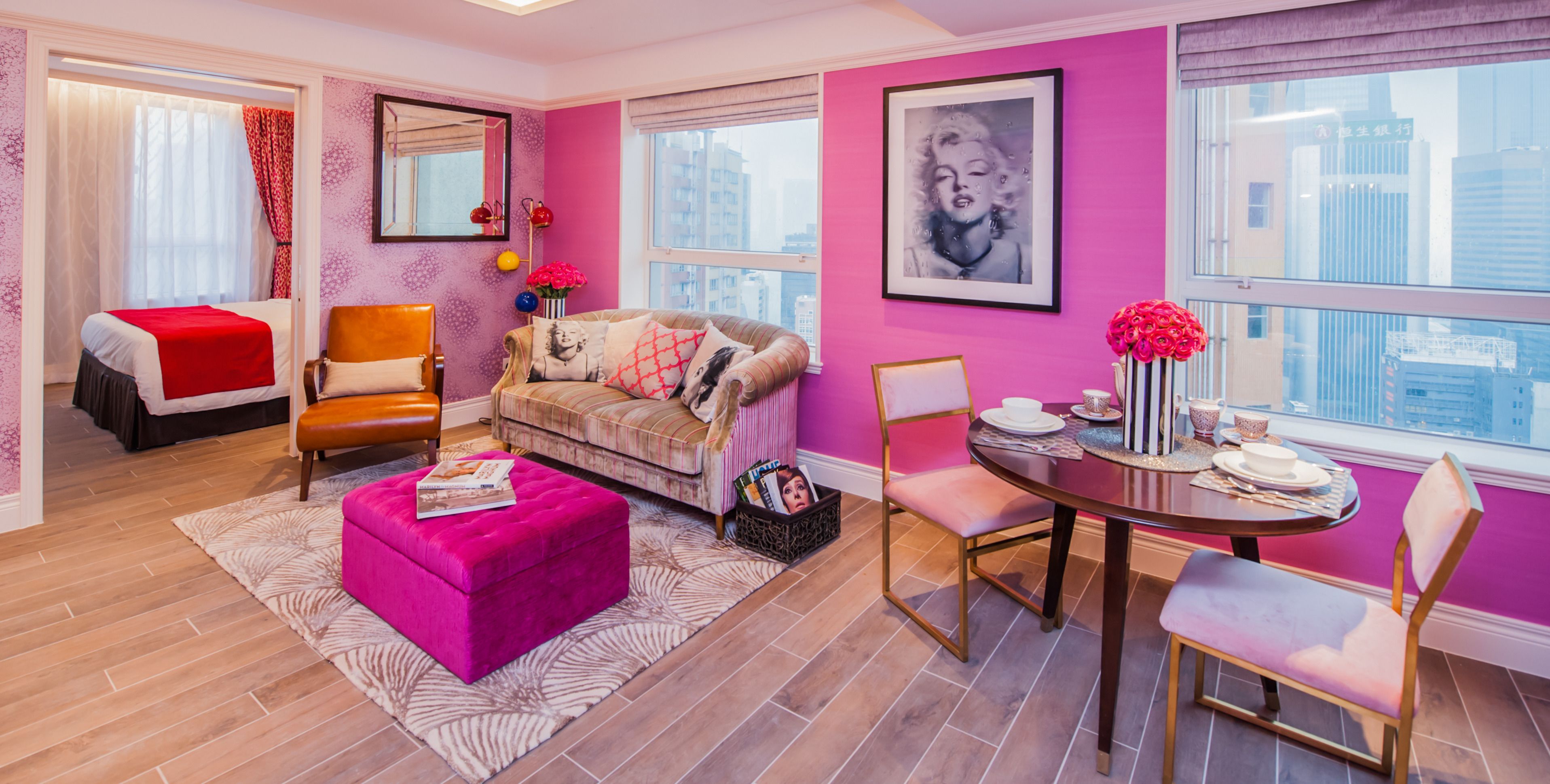 A modern living room with a pink wall, a large window, a sofa, a coffee table, and a dining area with chairs and a table set for two.