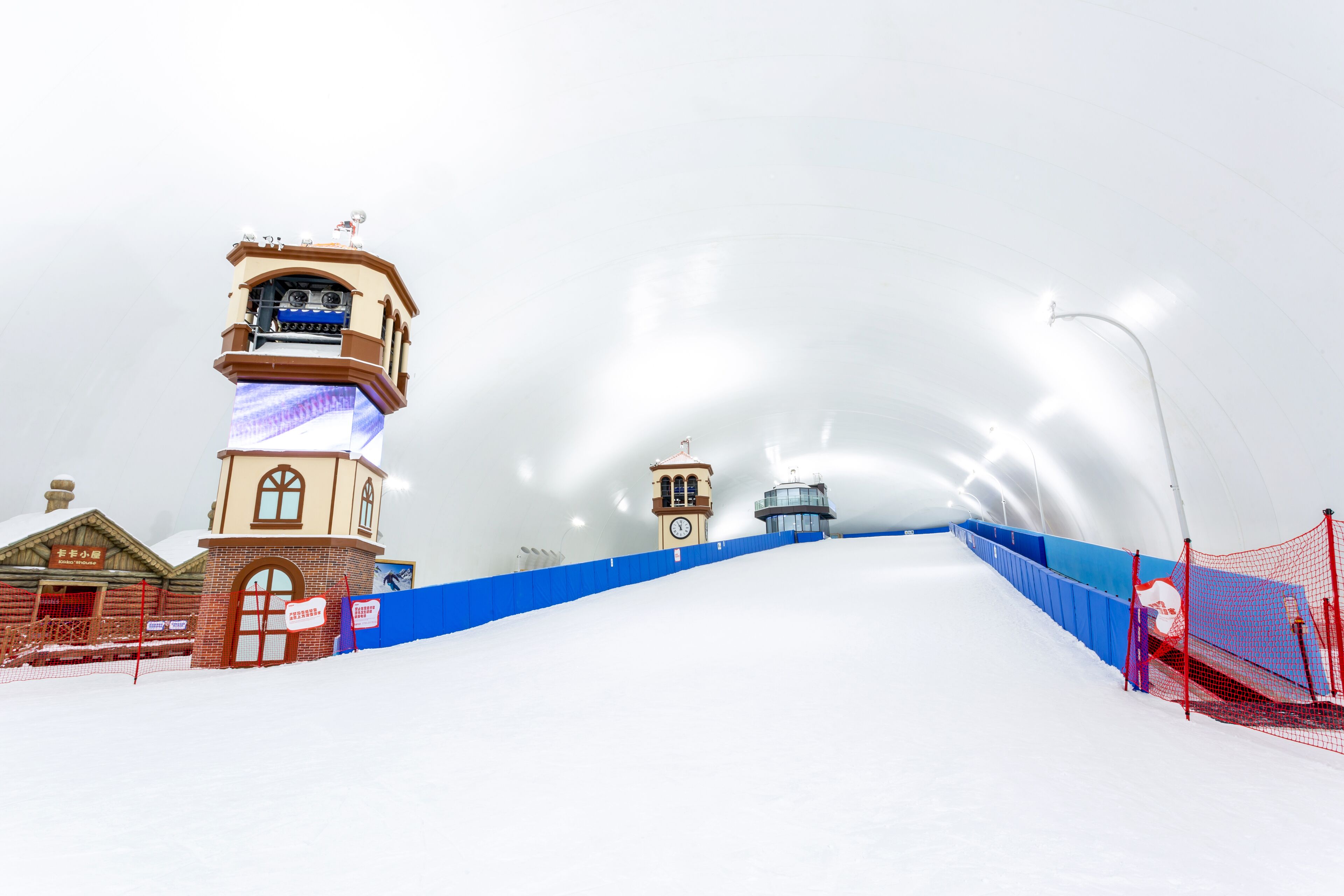 Pista de esquí nevada con una torre de control, una torre del reloj y una barrera de seguridad azul.