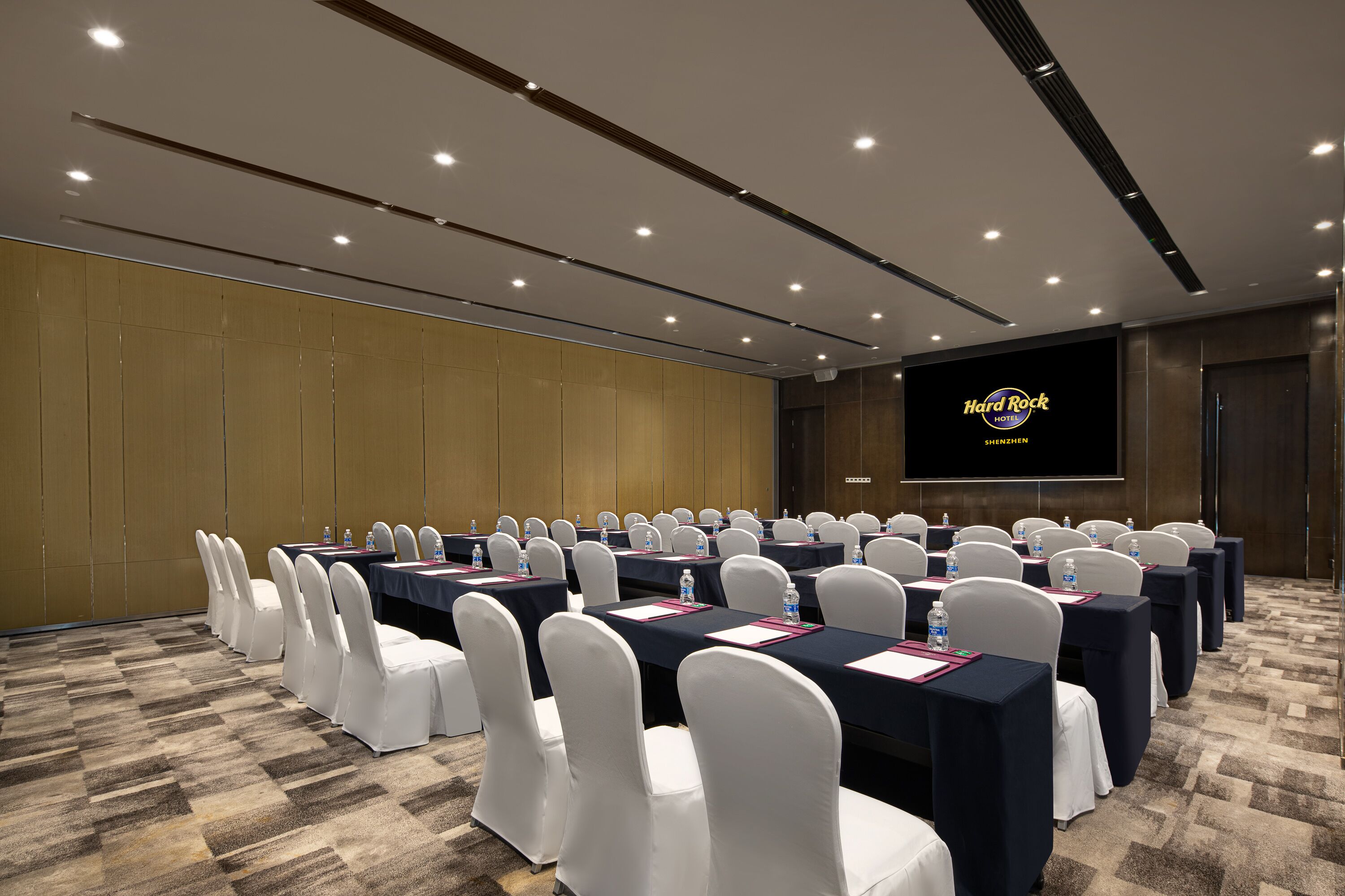 Een congrescentrum met rijen tafels en stoelen, een groot scherm waarop 'Hard Rock SHENZHEN' staat, en een vloerbedekking.