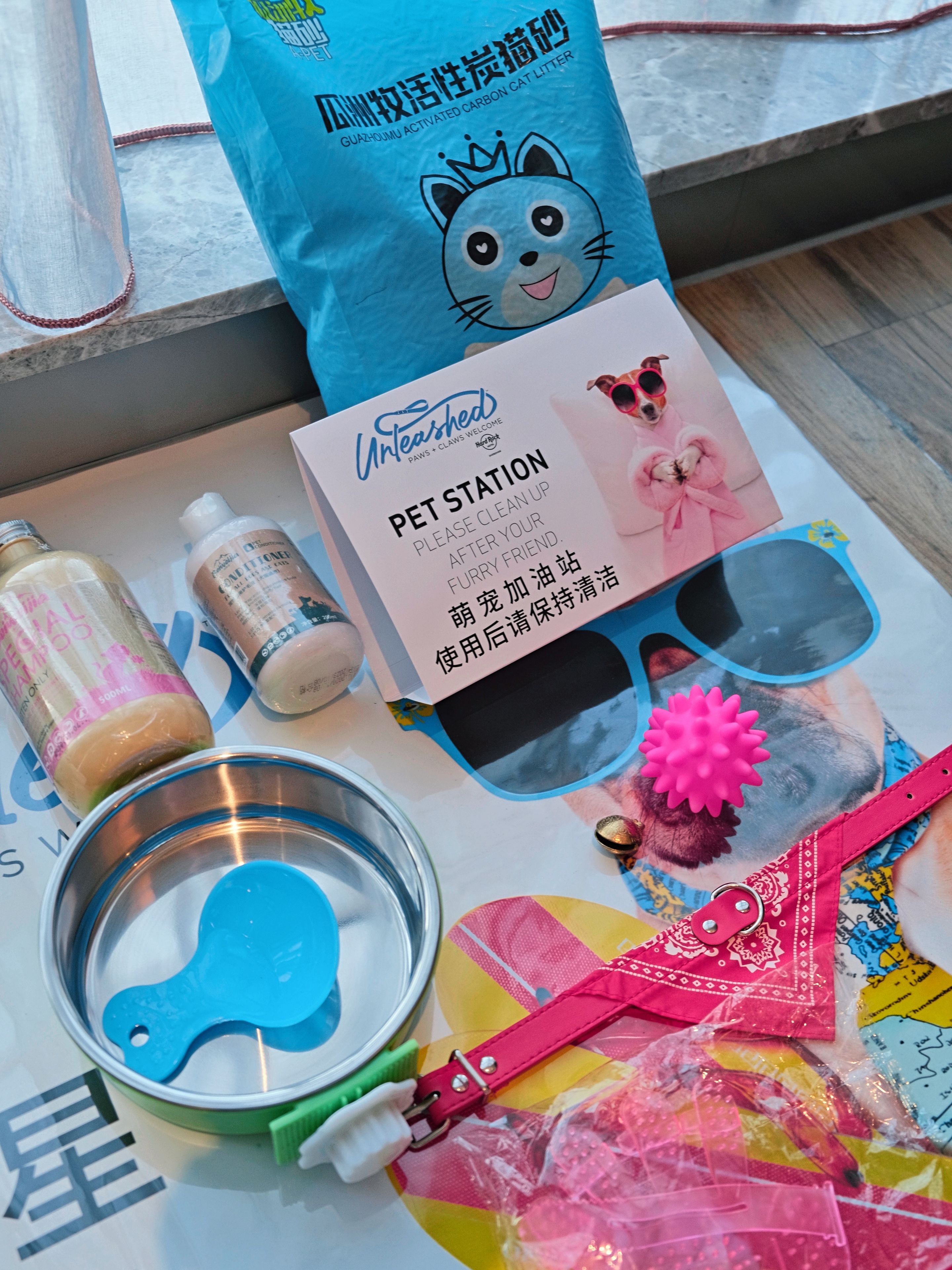 A pet station with a sign, a blue bag, a metal bowl, a bottle, a pair of sunglasses, and a pink toy.