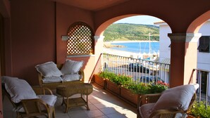 A balcony with wicker furniture, potted plants, and a view of the sea and boats.