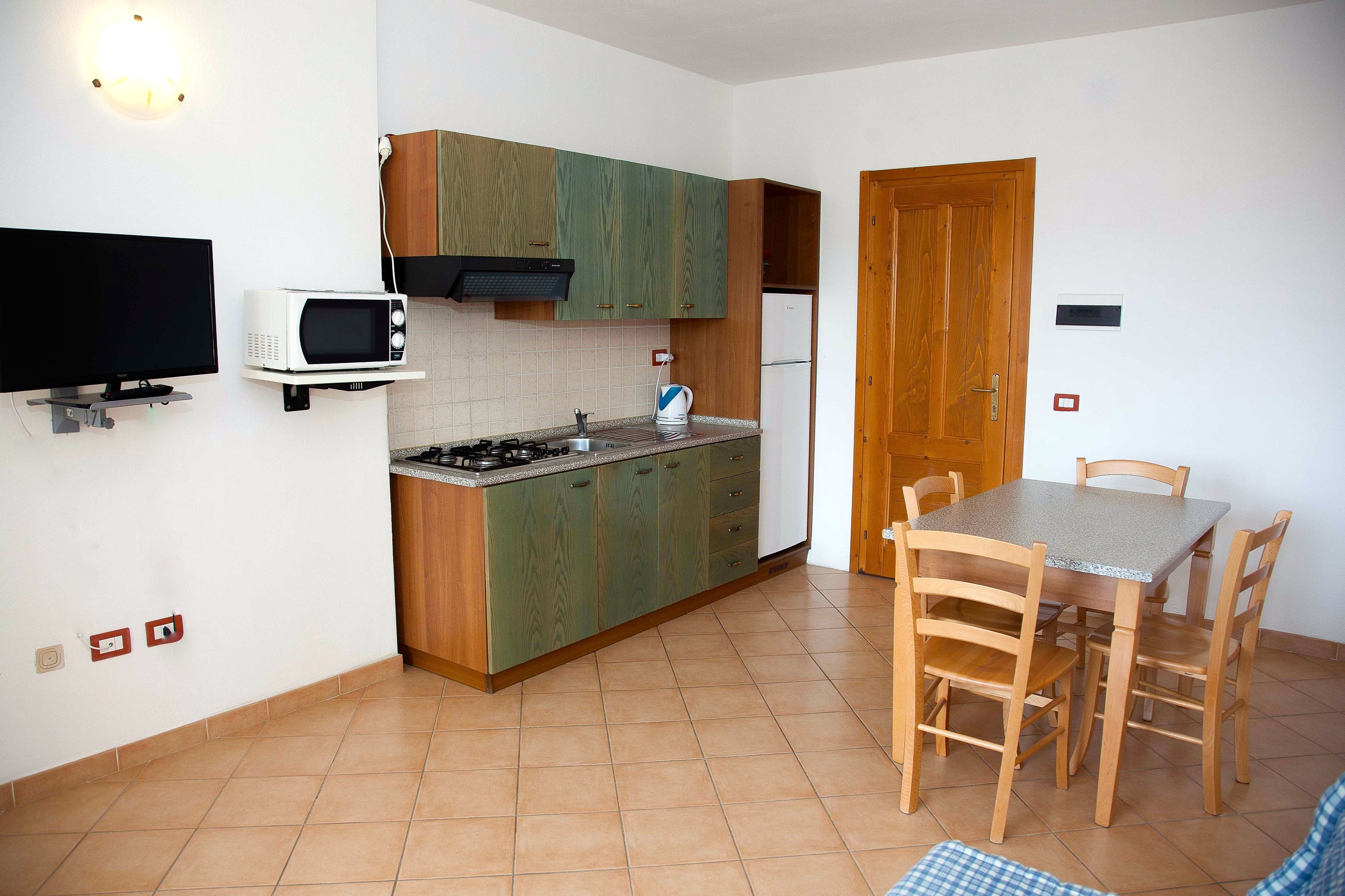 A compact kitchen with a microwave, stove, and wooden cabinets. There is a dining table with chairs and a television mounted on the wall.