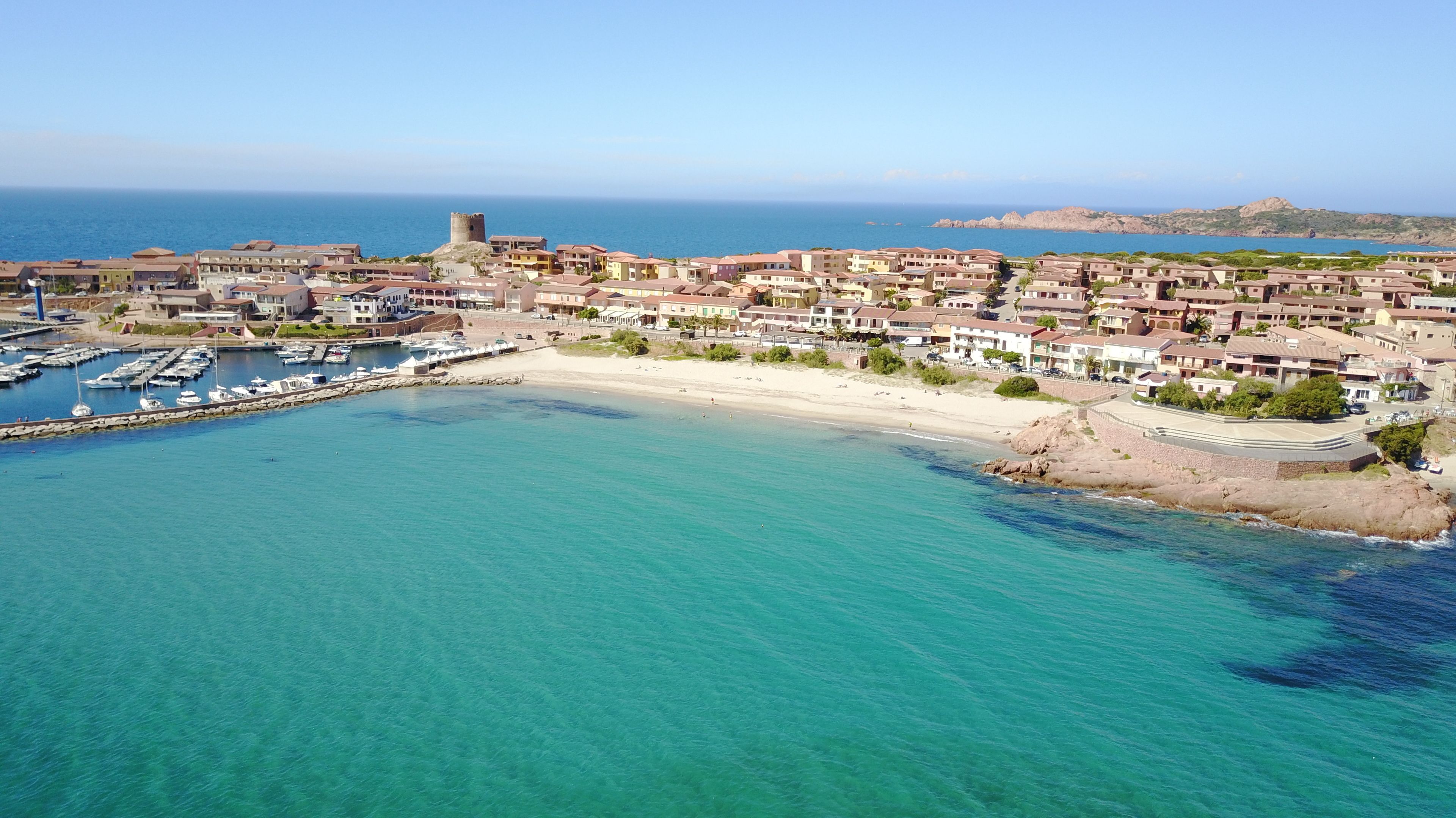 Vista aérea de un pueblo costero con puerto deportivo, playa de arena y una torre histórica.