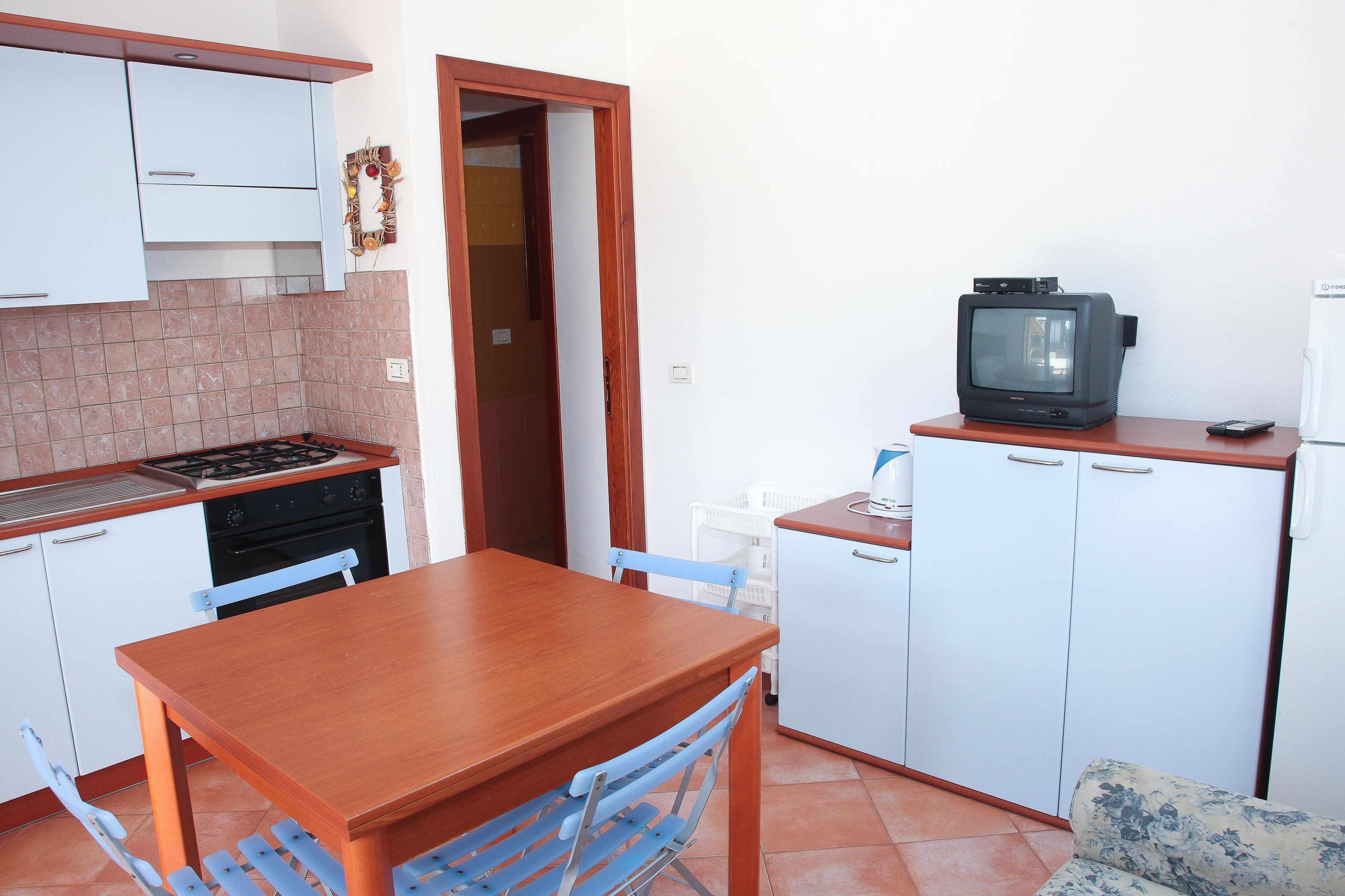 Een kleine keuken met een houten tafel en stoelen, een kleine televisie en eenvoudige apparaten.