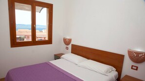 A bedroom with a bed, a nightstand, a wall-mounted lamp, and a window with a view of buildings and the sea.
