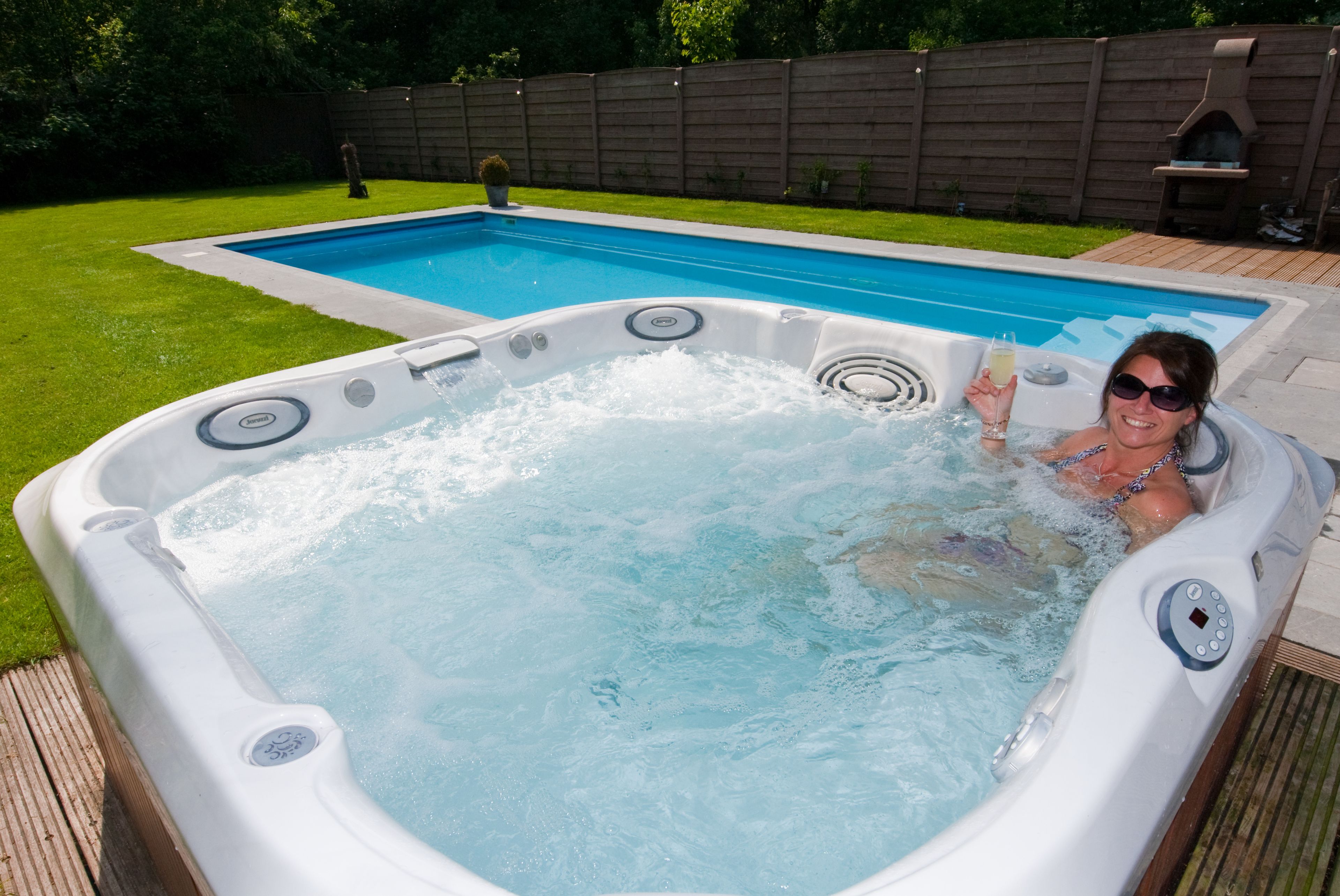 Een jacuzzi met daarin een persoon, omringd door een houten terras en een zwembad.