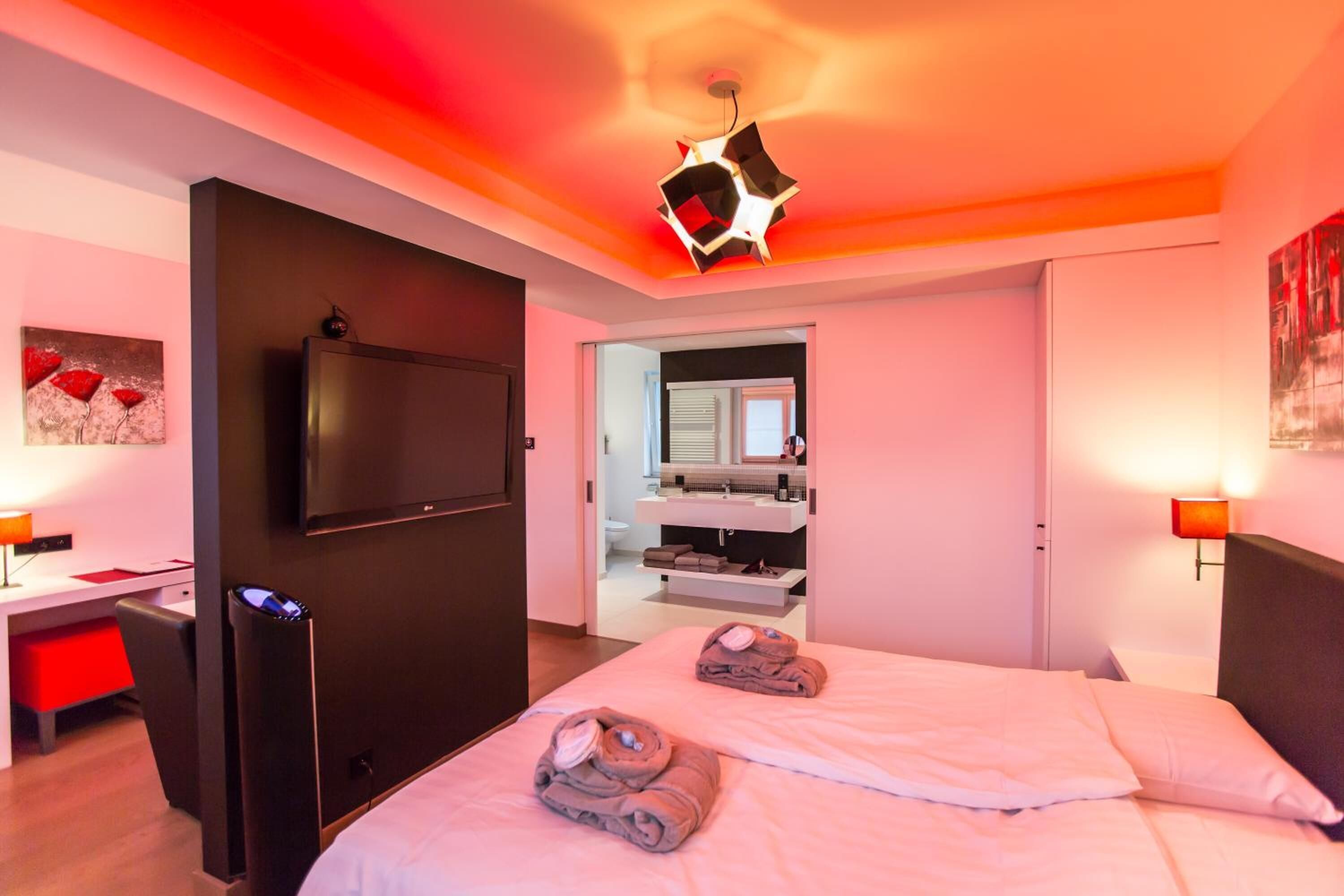 A modern hotel room with a bed, bedside tables, a TV, and a bathroom visible through an open door.