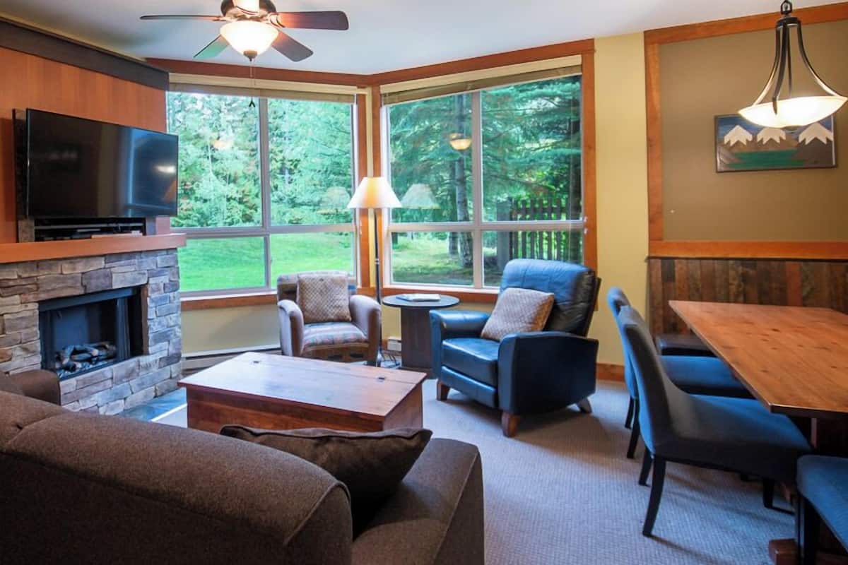 A cozy living room with a stone fireplace, comfortable seating, and large windows offering a view of the outdoors.