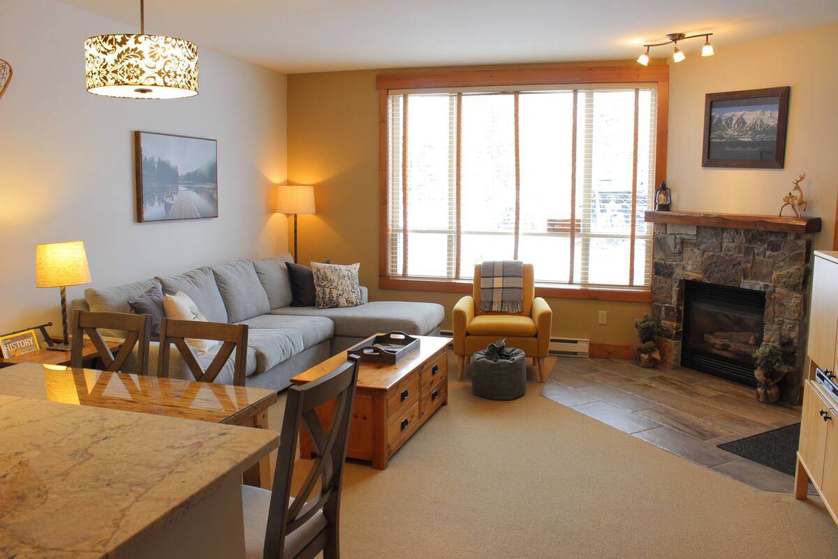 A cozy living room with a fireplace, a sofa, a dining table, and a large window.