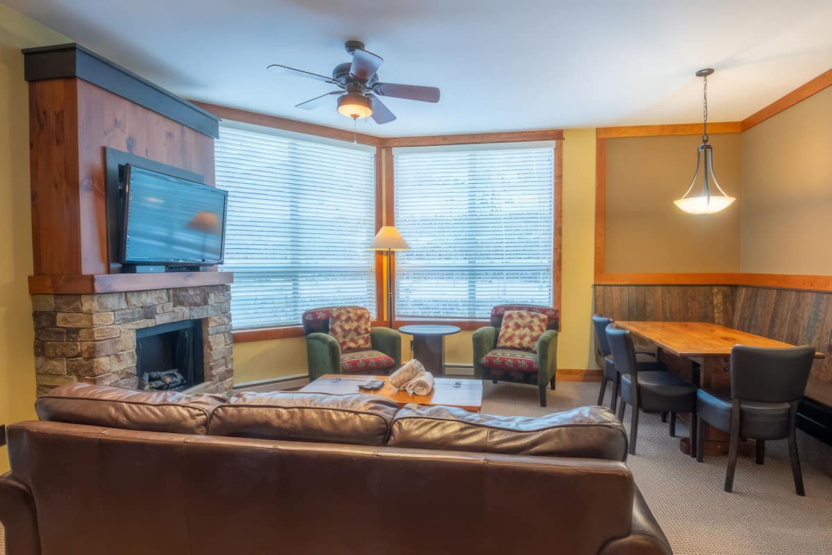 A cozy living area with a stone fireplace, leather sofa, and a television. There are two chairs, a small table, and a wooden bench.
