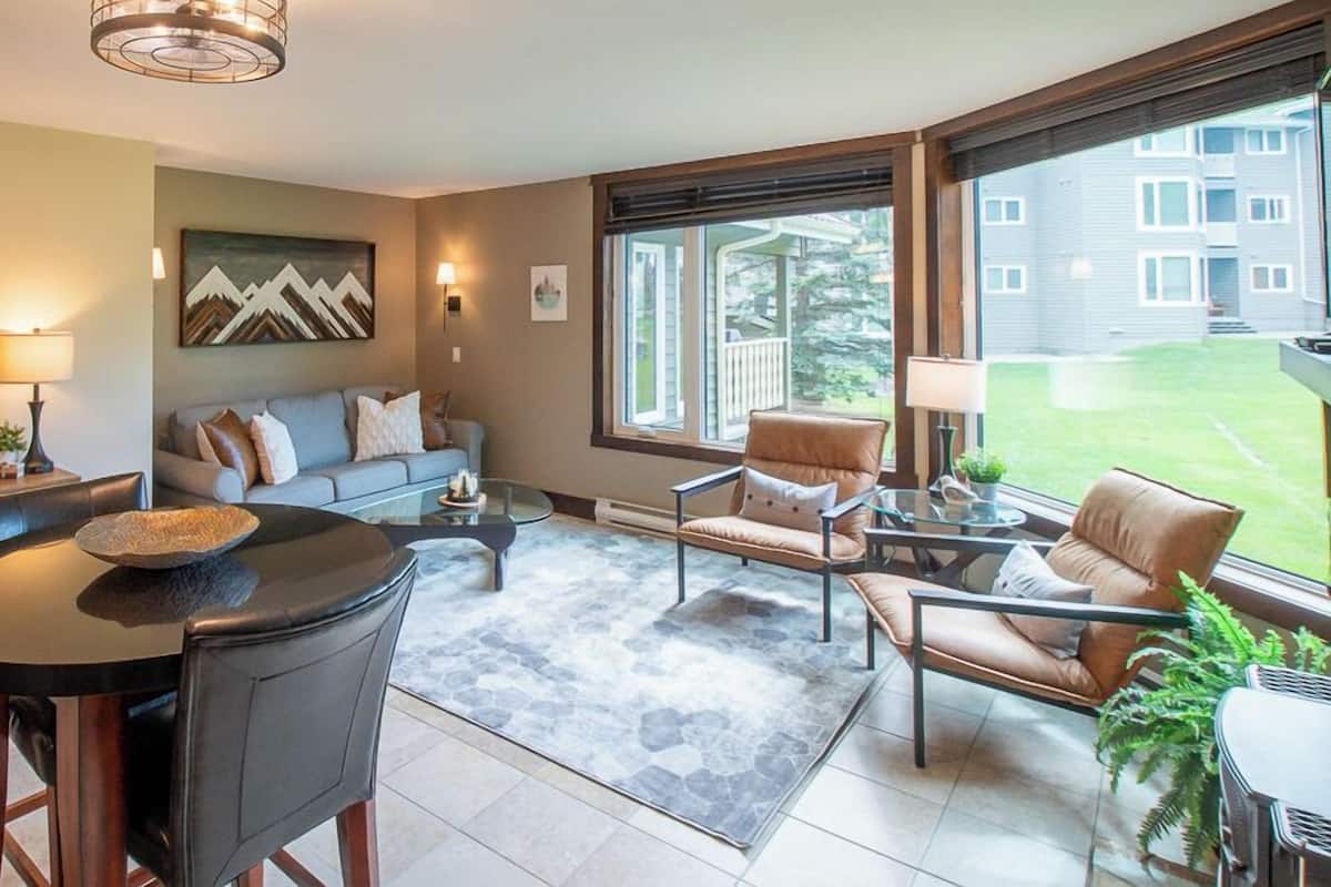 A modern living room with a sofa, two armchairs, a coffee table, and a dining table. There is a large window providing a view of the outside.