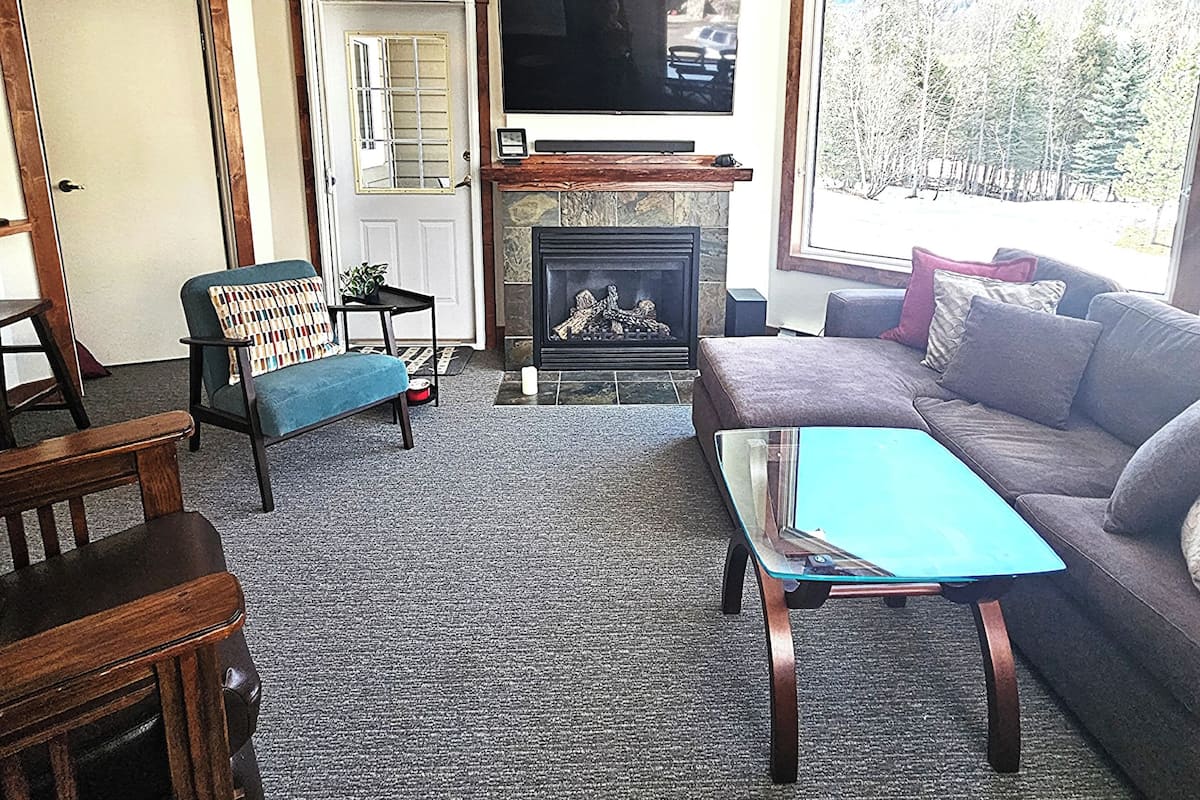 A living room with a fireplace, a television mounted above it, a ceiling fan, and a large window offering a view of trees.