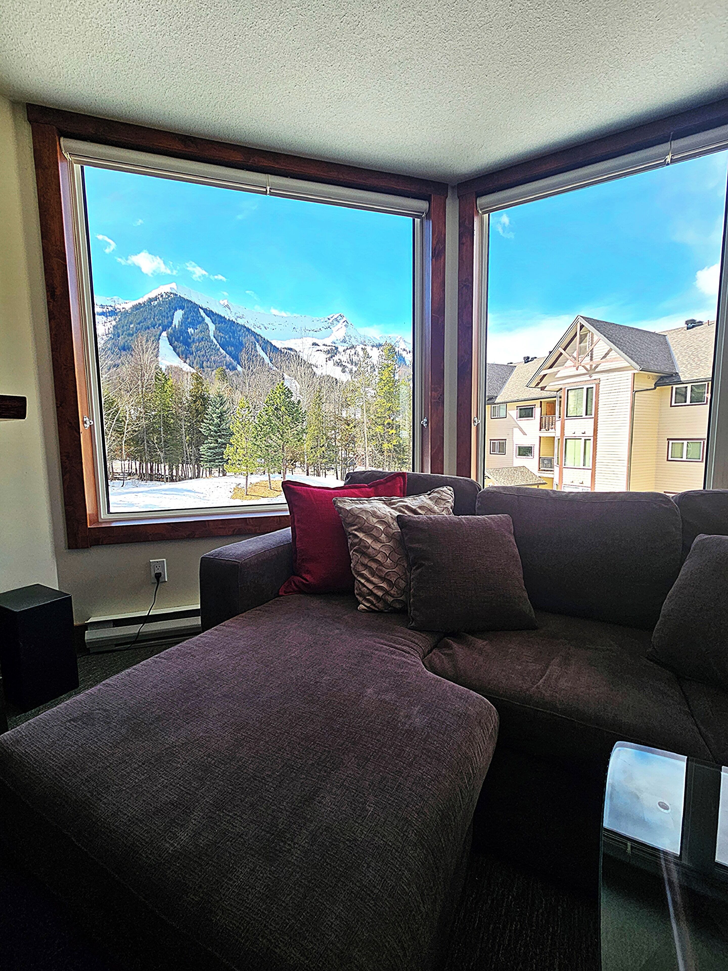 A room with a large window offering a mountain view, a dark-colored sofa, and a red pillow.