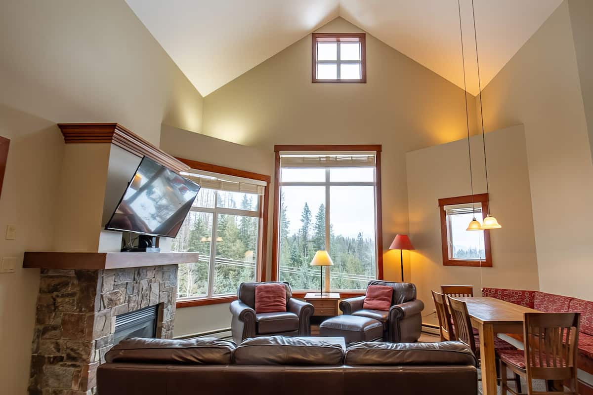 A spacious living room with a stone fireplace, leather sofas, a dining table, and a large window offering a view of trees.